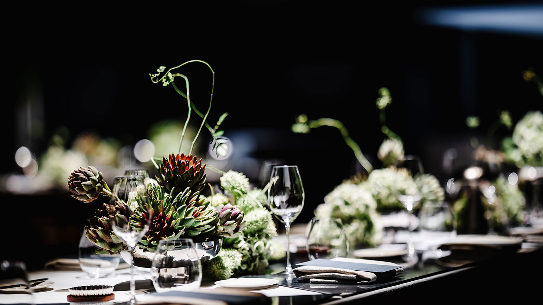 Een close-up van een elegante eettafel gedekt met wijnglazen, borden en servetten, met bloemstukken in het midden met groene en rode vetplanten, alles onder zachte verlichting.